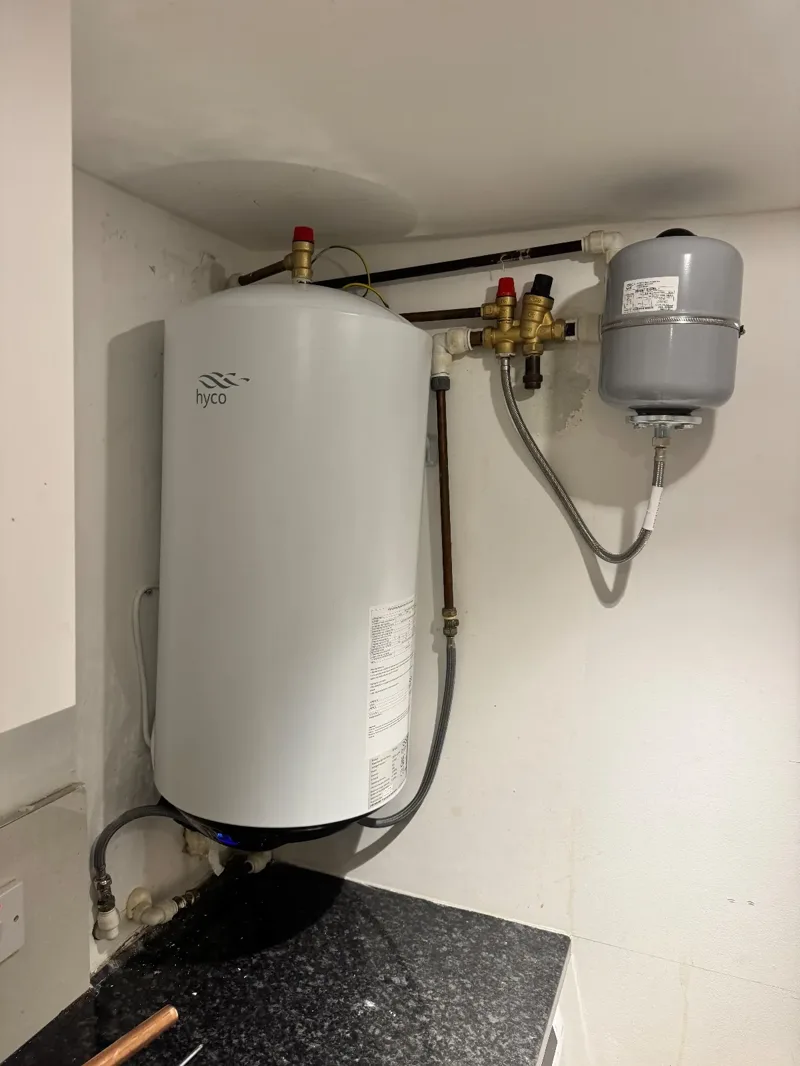 White wall-mounted hot water cylinder with its brass valve group and a grey expansion vessel on the wall beside it